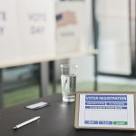 Tablet exibindo um cadastro eleitoral digital em uma estação de votação interna, com placas sobre o dia da votação.