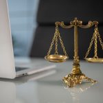 Balanças douradas da Justiça sobre uma mesa, ao lado de um laptop.