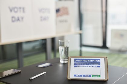 Tablet exibindo um cadastro eleitoral digital em uma estação de votação interna, com placas sobre o dia da votação.