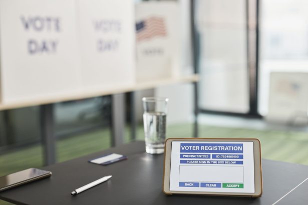 Tablet exibindo um cadastro eleitoral digital em uma estação de votação interna, com placas sobre o dia da votação.
